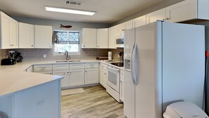 A modern kitchen with white cabinets, a stainless steel refrigerator, and a window with patterned curtains. - Island Mist B | The Beach is Your Backyard, Just Steps from the Sand! (Fort Walton Beach)