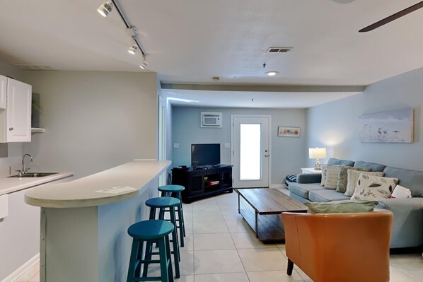 A modern kitchen with a bar area, a living room with a sofa and a TV, and a dining area with a wooden table.