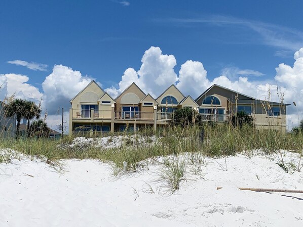 A beachfront property with multiple balconies and large windows, surrounded by sand and sparse vegetation.