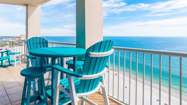 Een balkon met turquoise stoelen en een tafel, met uitzicht op een strand en de oceaan.