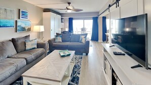 A modern living room with a sofa, coffee table, TV, and artwork on the wall. - Regency Towers #203 West | Beach + Pool Access! (Pensacola Beach)