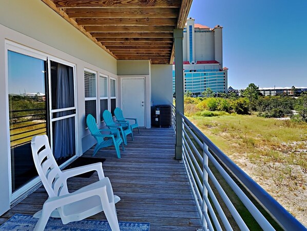 A covered outdoor area with a wooden deck, white plastic chairs, and a view of a building and landscape. - Lakefront Condo, Close to Beach with Resort Amenities (Orange Beach)