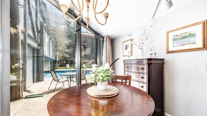 A dining area with a wooden table, chairs, a vase of flowers, and a view of a swimming pool through large glass doors.