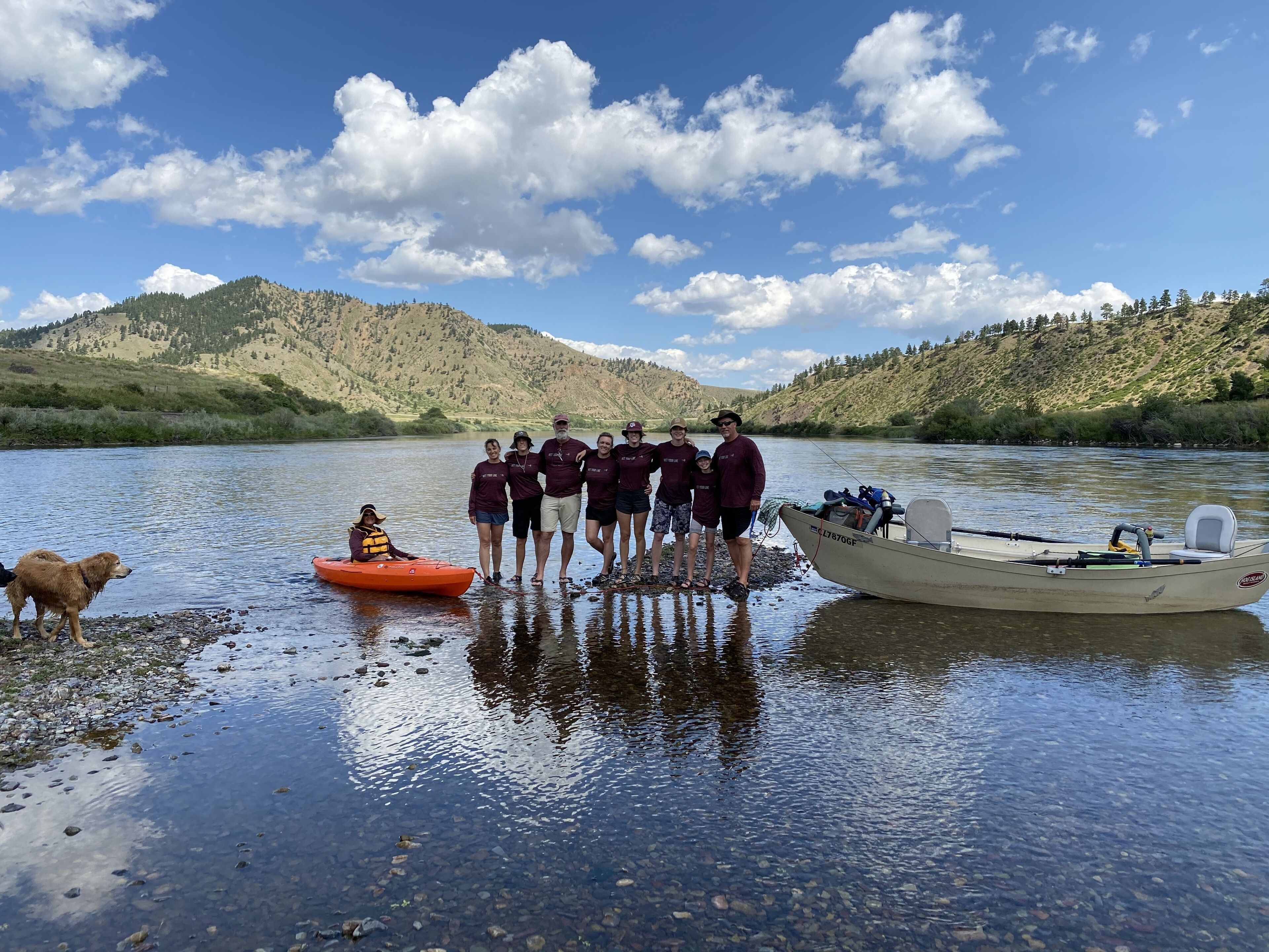 A group of people standing by a river with boats and a dog.