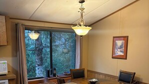 A dining area with a wooden table and chairs, a pendant light, a window with curtains, and a framed picture on the wall. - Cozy Cabin in the Mountains (Newland)