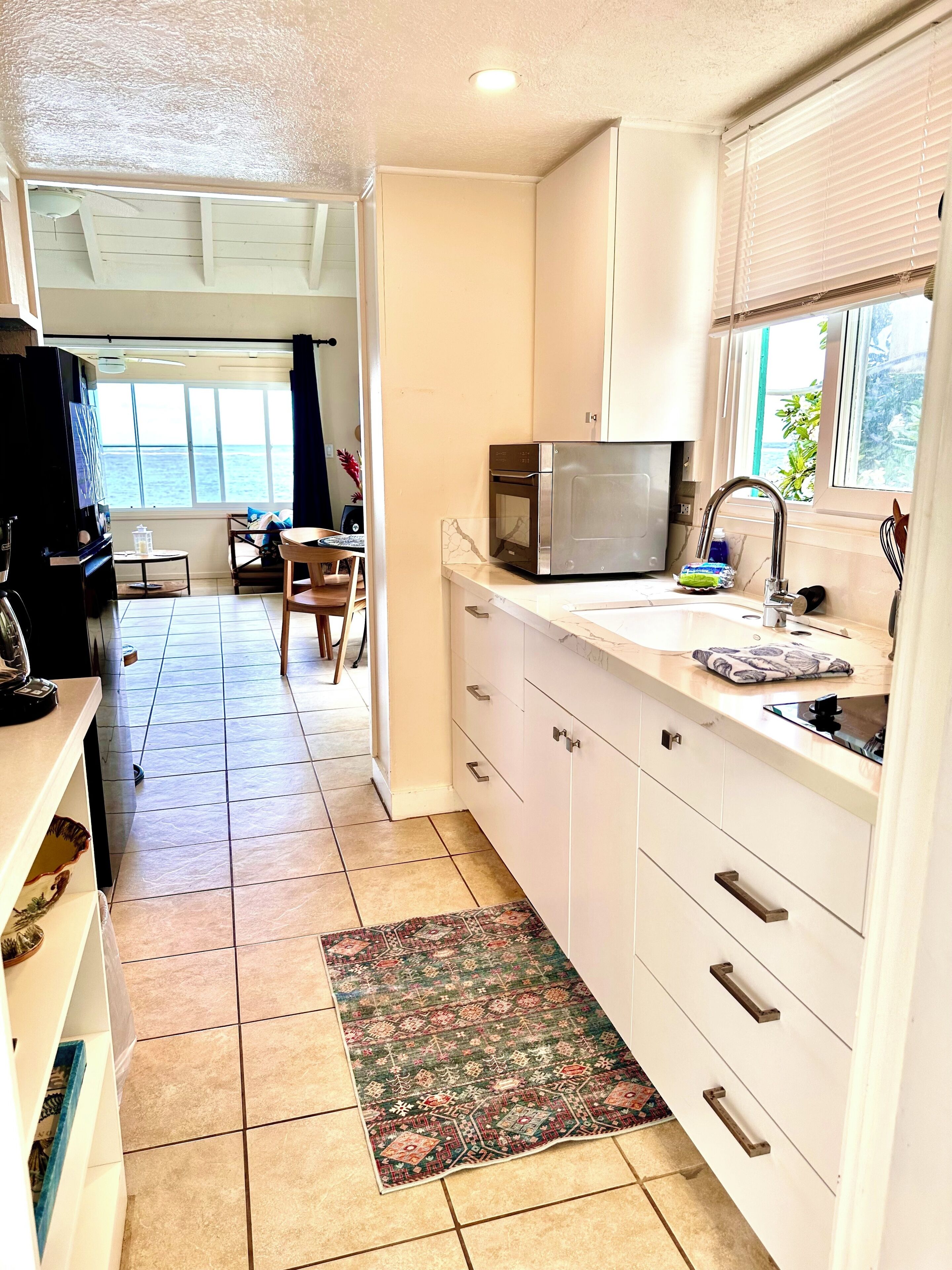 A compact kitchen with white cabinets, a microwave, and a sink. A patterned rug is on the floor, and there's a view of the sea through a window.