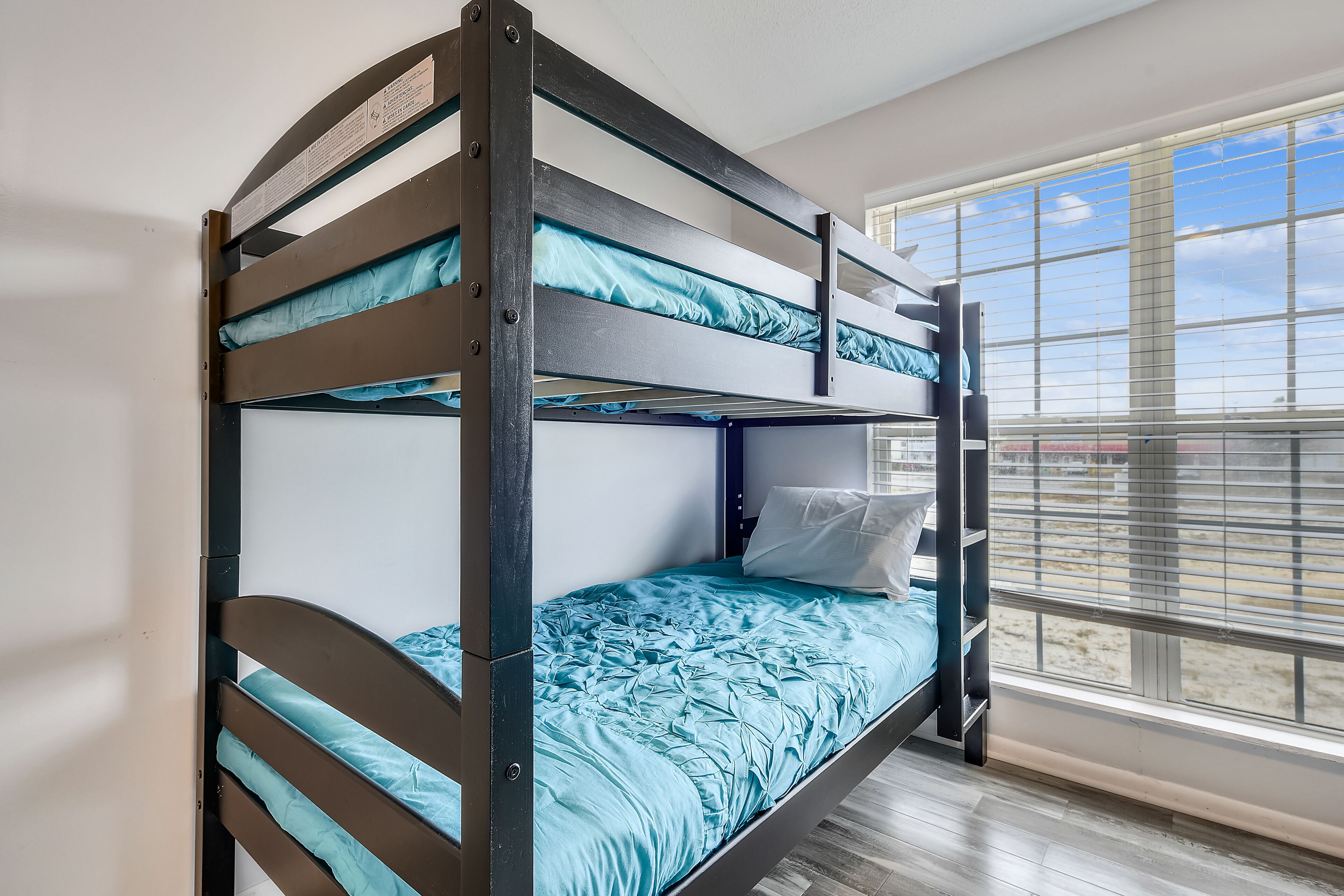 A bunk bed with a blue mattress and pillows, a window with blinds, and a wooden floor.
