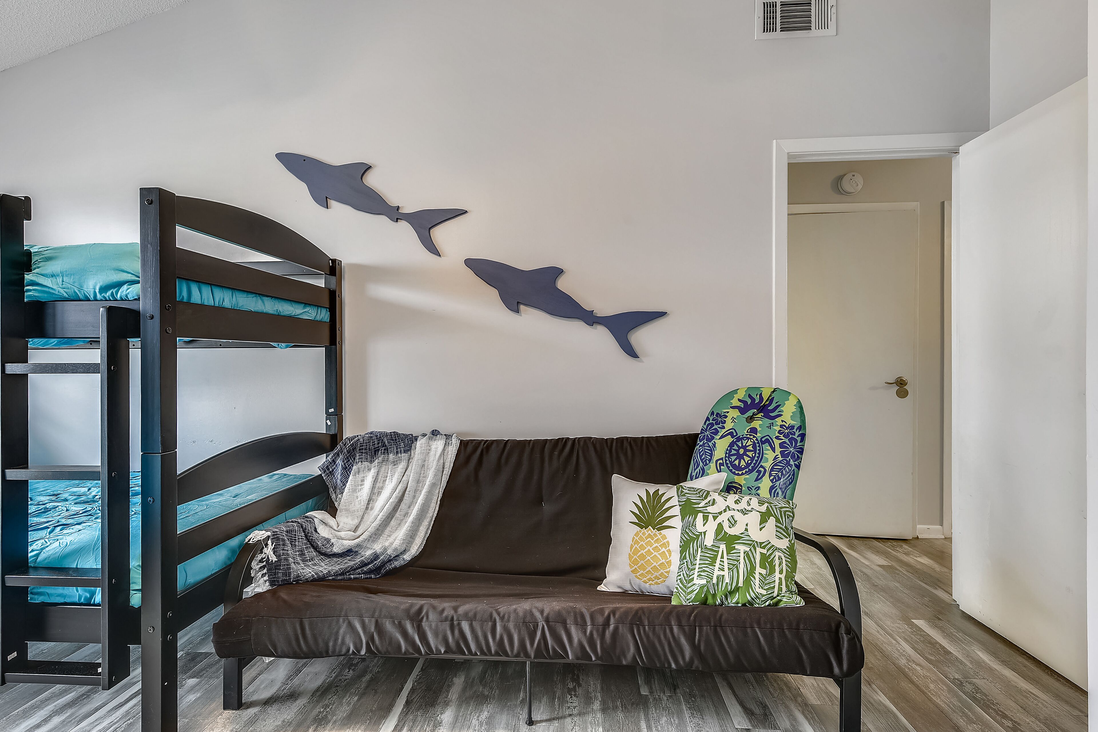 A bunk bed with a blue mattress, a brown sofa with a pineapple pillow, and two shark wall decals.
