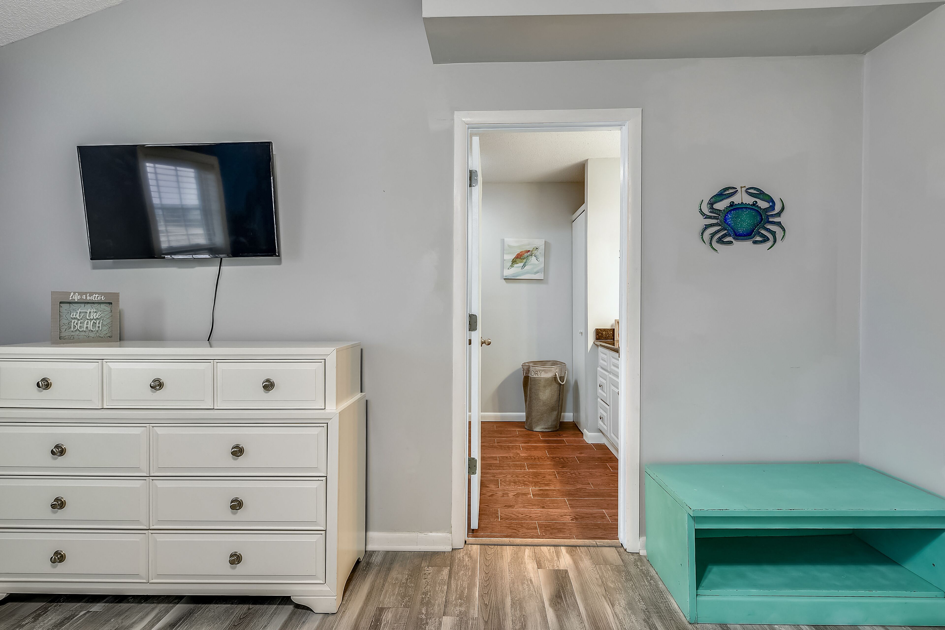 A room with a white dresser, a teal bench, a television, and a door leading to another room.