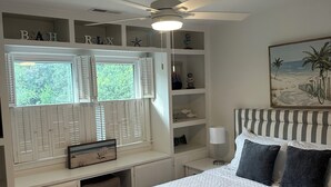 Ein Schlafzimmer mit einem Bett, einem Deckenventilator, einem Fenster mit Rollläden und einem Strandbild.