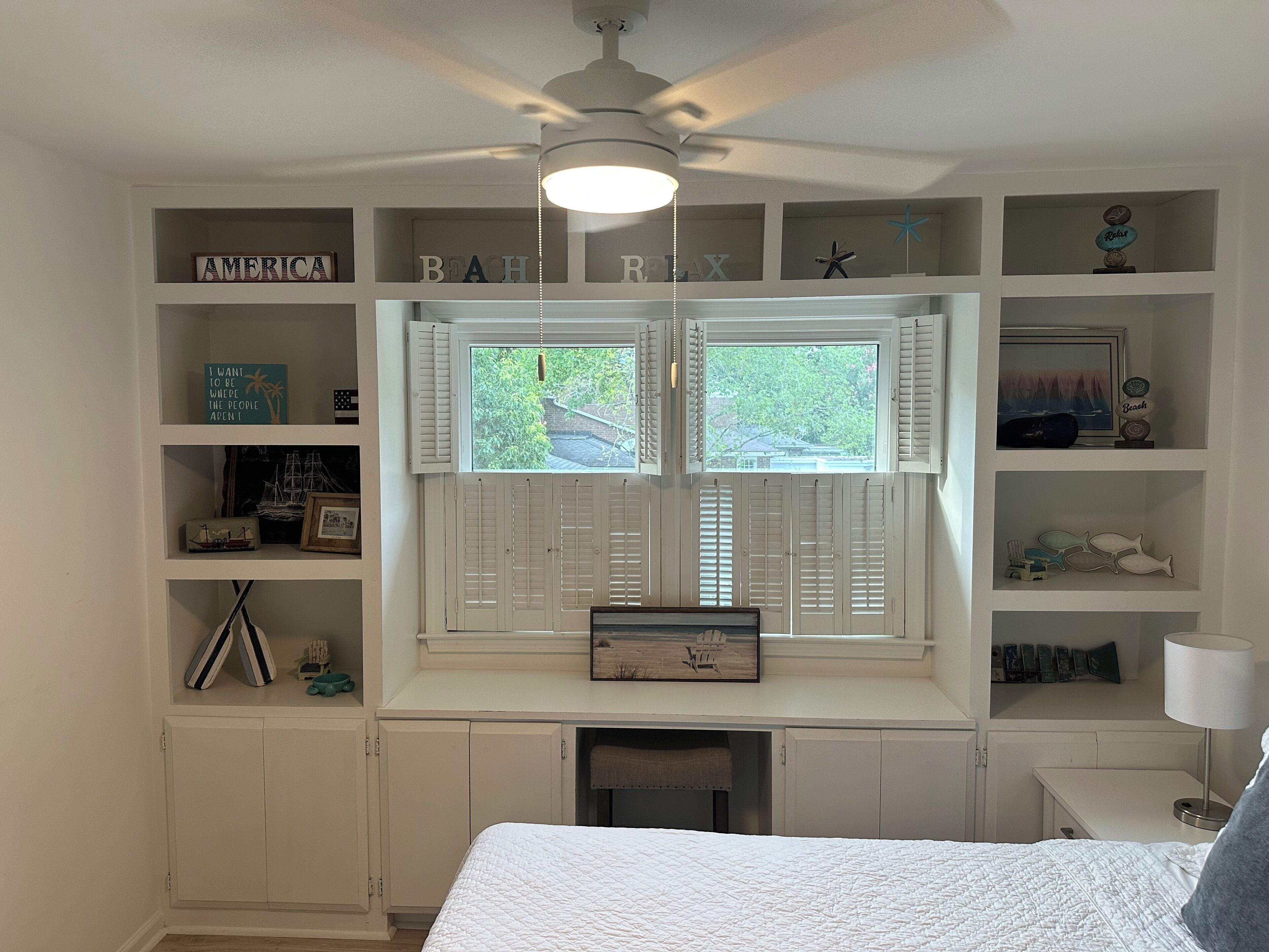 A bedroom with a large bed, a desk, and a window with shutters. Shelves with various items and a ceiling fan are also visible.