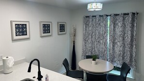 Eine moderne Küche mit Spüle, Armatur und einem kleinen Tisch mit Stühlen. An der Wand hängen drei gerahmte Kunstwerke und ein Blumenvasen mit Trockenpflanzen.