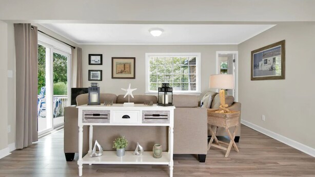 A living room with a beige sofa, a white console table, and a lamp. There are framed pictures on the walls and a window with blinds. - Tara Cove Cottage (Ogunquit)