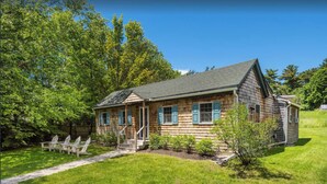 A wooden cabin with a porch, surrounded by a grassy yard and trees. - Tara Cove Cottage (Ogunquit)