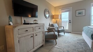 Une chambre à coucher avec un grand téléviseur à écran plat fixé au mur, une commode en bois, deux fauteuils, un lit recouvert d’une couverture bleue, une fenêtre donnant sur un paysage agréable et une porte.
