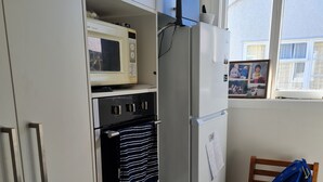 A compact kitchen with a microwave, oven, and refrigerator. A wooden chair with a blue bag is visible. - Womans Beauty  (Christchurch)