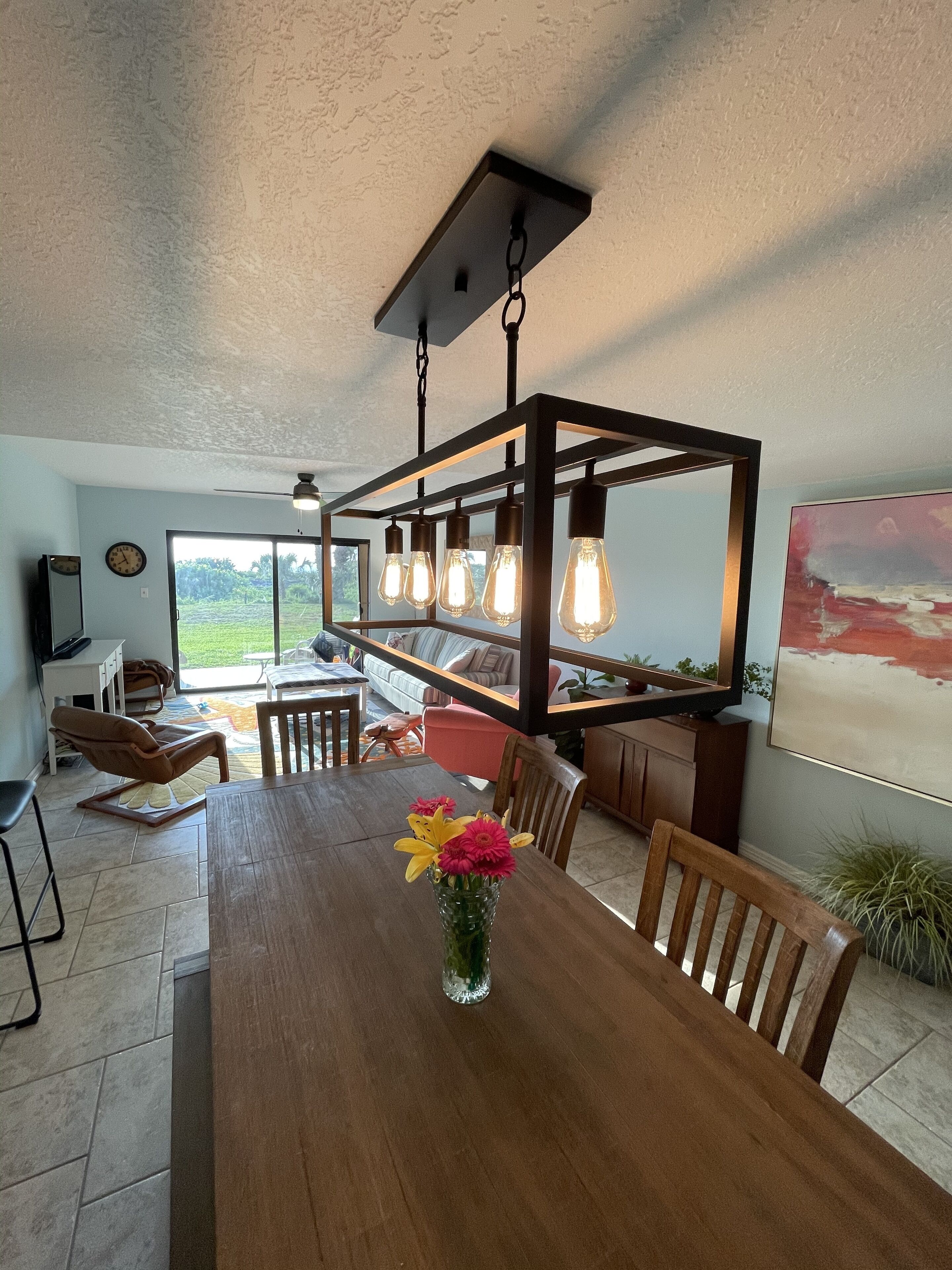 A dining area with a wooden table, chairs, a vase with flowers, and a hanging light fixture.