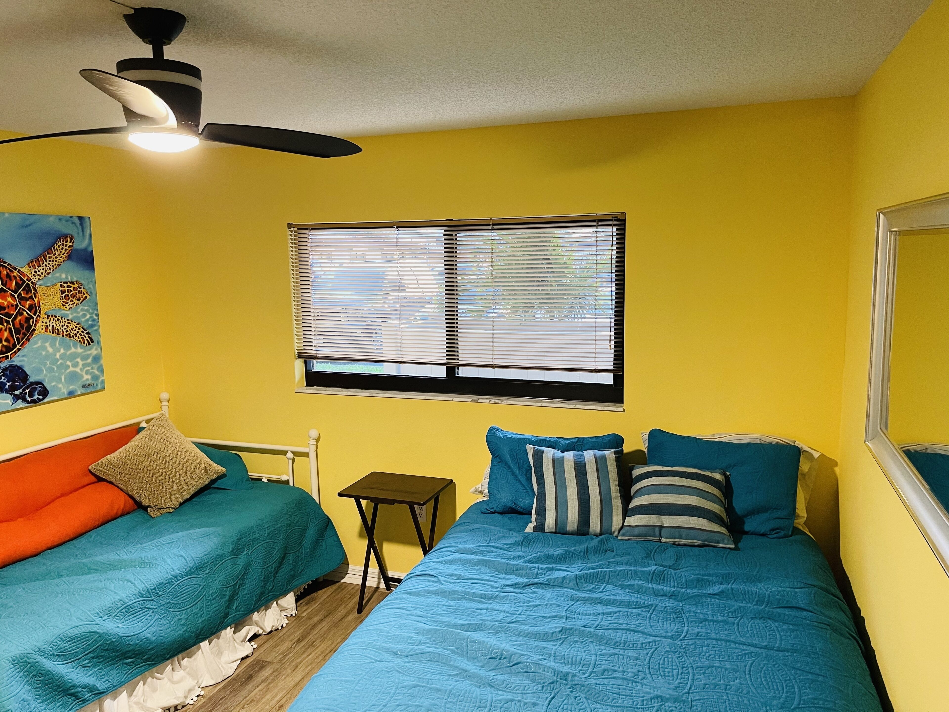A room with two beds, a ceiling fan, a window with blinds, and a painting on the wall.