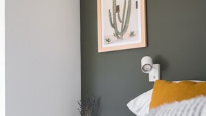 Une chambre à coucher avec un lit, une œuvre d’art représentant un cactus encadré, une petite plante en pot et un luminaire fixé au mur.