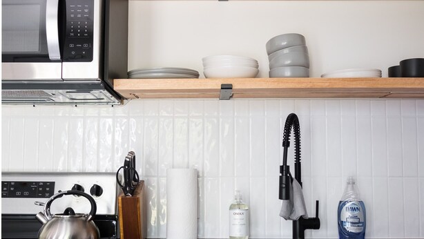 Une cuisine moderne équipée d’un four à micro-ondes, d’une bouilloire et d’appareils électroménagers en acier inoxydable. Des étagères en bois, situées au-dessus du plan de travail, servent à ranger la vaisselle et une plante d’ornement y est posée.