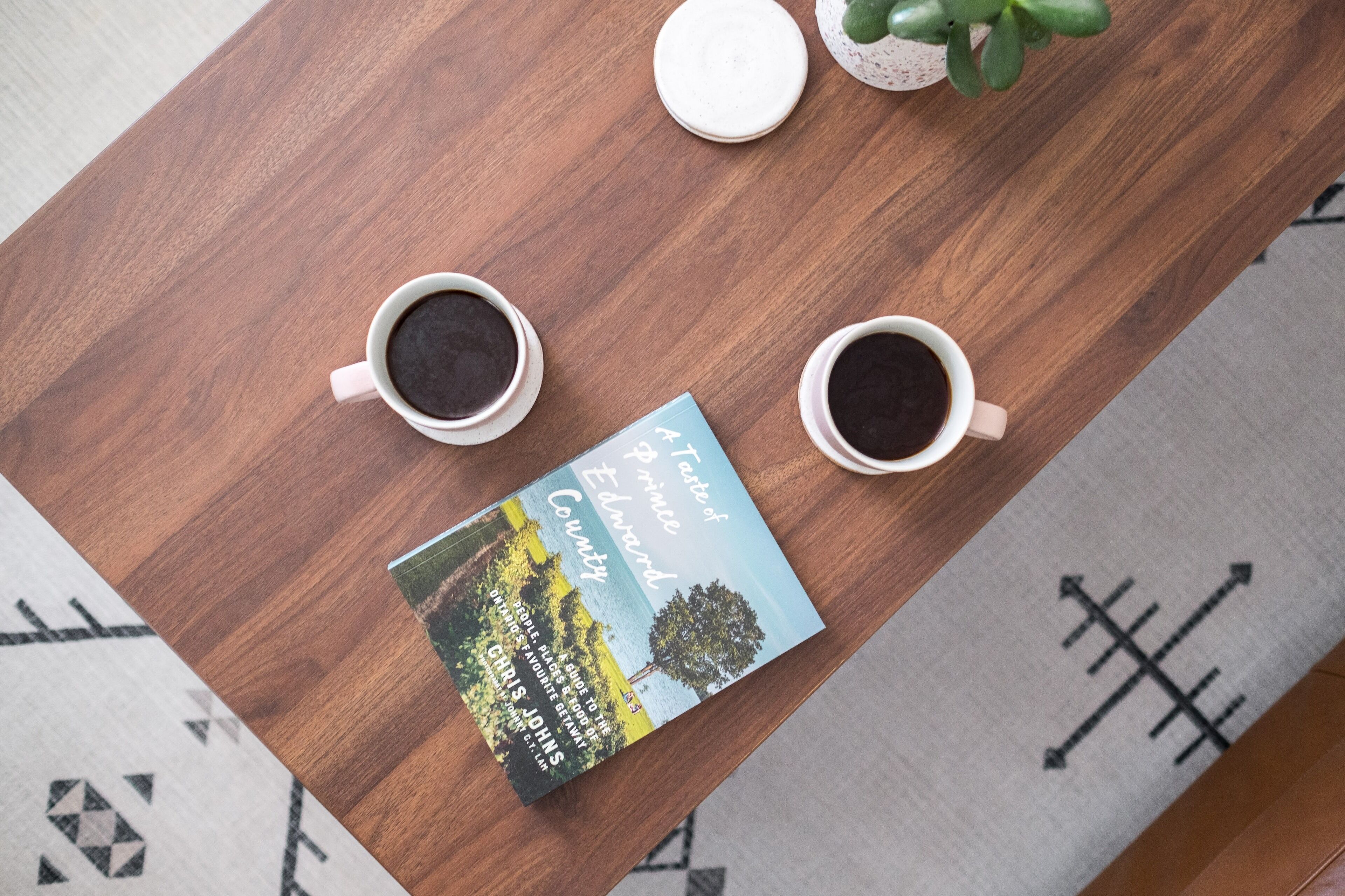 Une table en bois sur laquelle reposent deux tasses de café, un livre intitulé « Saveurs de la région de Prince-Édouard » et une petite plante en pot.