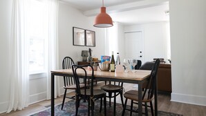 Un coin repas avec une table en bois, des chaises noires, un luminaire suspendu rouge et une vue sur le salon à travers une porte ouverte.