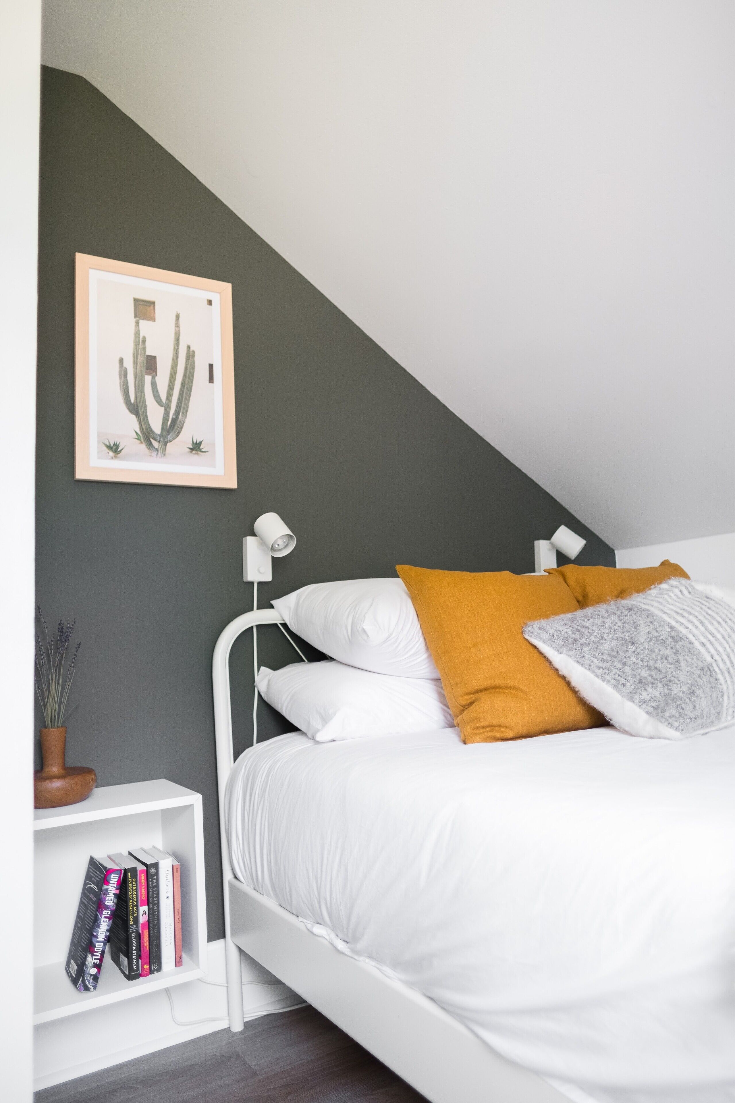 A bedroom with a white bed, a nightstand with a lamp, a framed cactus artwork, and a bookshelf with books.
