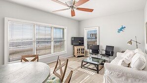 A spacious living room with a ceiling fan, large windows, a TV, a white sofa, and a dining area with a table and chairs. - Stay @ Ocean Spray at the Blue Horizon (Sullivan's Island)
