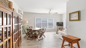 A dining area with a wooden table and chairs, a television, and a window with blinds. - Stay @ Ocean Spray at the Blue Horizon (Sullivan's Island)