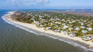 An aerial view of a coastal residential area with houses along the beach, clear skies, and a distant view of a city skyline. - Stay @ Ocean Spray at the Blue Horizon (Sullivan's Island)