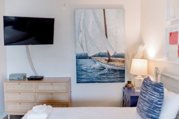 A bedroom with a bed, a dresser, a painting of a sailboat, a television mounted on the wall, and a nightstand with a lamp and a decorative item.