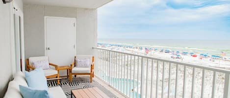 A balcony with a view of the beach, a pool, and a seating area with a table and chairs.