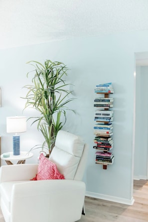 A room with a white chair, a tall plant, and a wall-mounted bookcase.