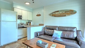 A kitchen with white cabinets, a refrigerator, and a microwave. There is a brown leather couch with a pillow that says "The Beach is my Happy Place." A wooden coffee table with a basket and a magazine is in front of the couch. - Ocean front condo with amazing ocean views! (Hilton Head Island)