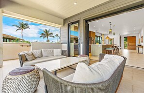 A modern living area with a sofa, chairs, and a table. There is a view of a balcony with palm trees and a clear sky.