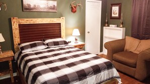A bedroom with a wooden bed, a nightstand, a lamp, a chair, a door, and a shelf with items.