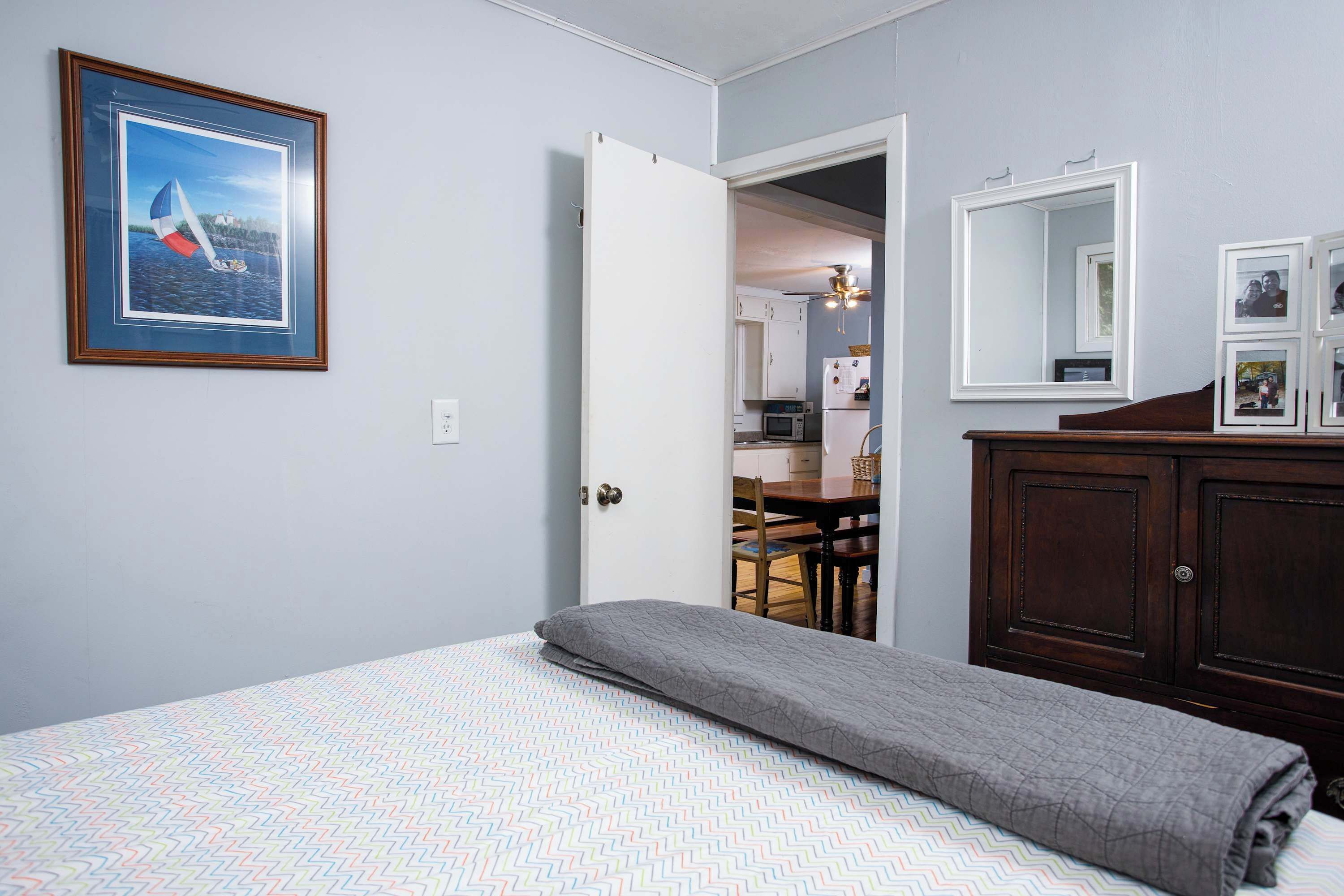 Quarto com cama, manta cinzenta, quadro emoldurado, cômoda de madeira e porta que dá para uma cozinha.