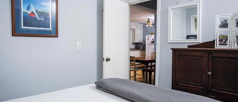 Quarto com cama, manta cinzenta, quadro emoldurado, cômoda de madeira e porta que dá para uma cozinha.
