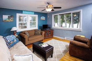 A living room with a ceiling fan, a television, a sofa, a coffee table, and a window with a view of trees.
