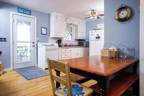 A kitchen with white cabinets, a wooden dining table, a chair with a floral seat, a clock on the wall, and a door with a sign.