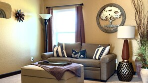 Un salon avec un ventilateur de plafond, un canapé, un pouf, une lampe et une œuvre d’art décorative représentant un arbre sur le mur.