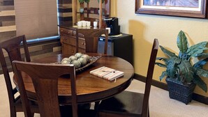 Un coin repas avec une table ronde en bois, des chaises, un lustre, une horloge, un tableau encadré et une plante en pot.