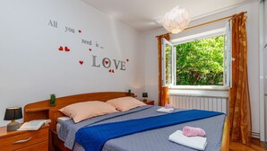 Ein ordentlich bezogenes Bett mit blauer Decke, eine Wand mit dekorativer Liebesmotive und ein Fenster mit Vorhängen.