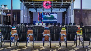 An outdoor seating area with black chairs and a large screen displaying a surfing event. - Bella Bonita by AvantStay | Pool, Peloton & Bikes, Golf Cart (Santa Rosa Beach)
