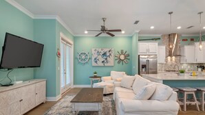 A modern living room with a white sofa, a coffee table, and a TV on a cabinet. There is a kitchen area with a fridge, stove, and hanging lights. - Bella Bonita by AvantStay | Pool, Peloton & Bikes, Golf Cart (Santa Rosa Beach)