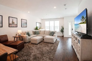 A modern living room with a sectional sofa, a flat-screen TV, and a wooden dining table.