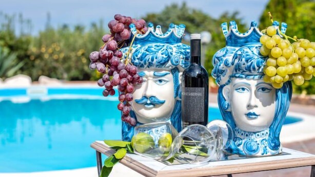 Dos estatuas de cerámica con uvas y una botella sobre una mesa junto a una piscina.