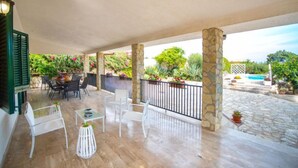 Un espacio al aire libre techado con pilares de piedra, una piscina y un patio con sillas y mesas blancas.