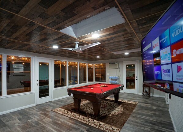 A pool table in a room with wooden ceilings and a large screen displaying streaming services.