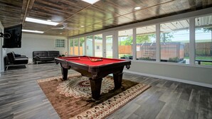 A pool table in a room with a wooden ceiling, large windows, and a view of the backyard.
